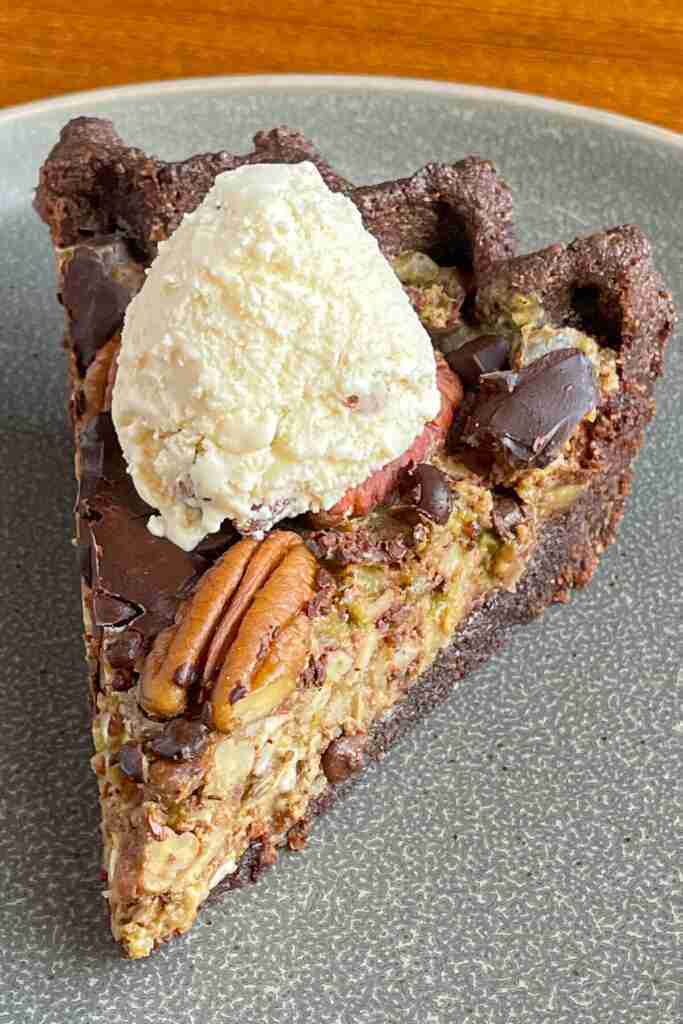 A slice of sugar-free, keto chocolate chip pecan pie in a chocolate pie crust and topped with a scoop of sugar-free butter-pecan ice cream.