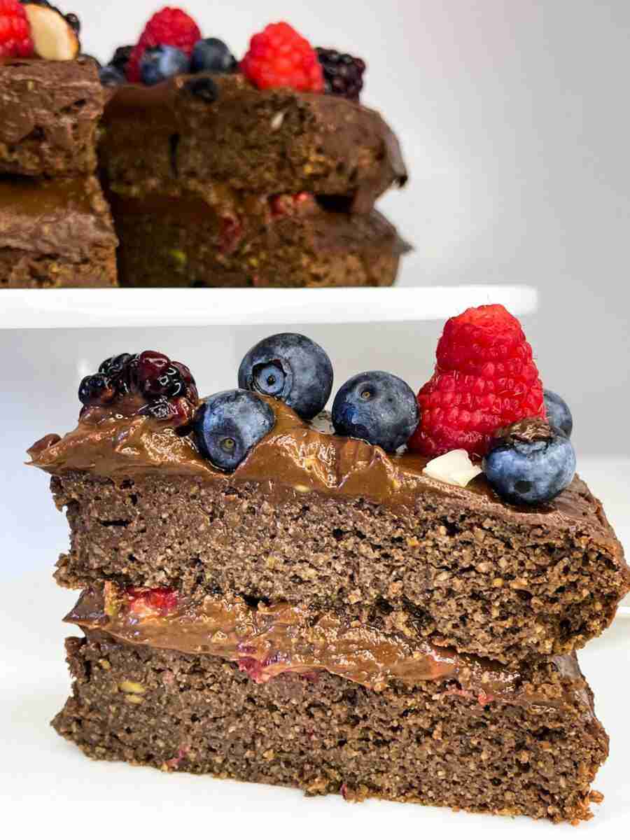 A slice of a sugarfree, glutenfree, keto chocolate raspberry layer cake topped with the cake behind it and topped with fresh berries.