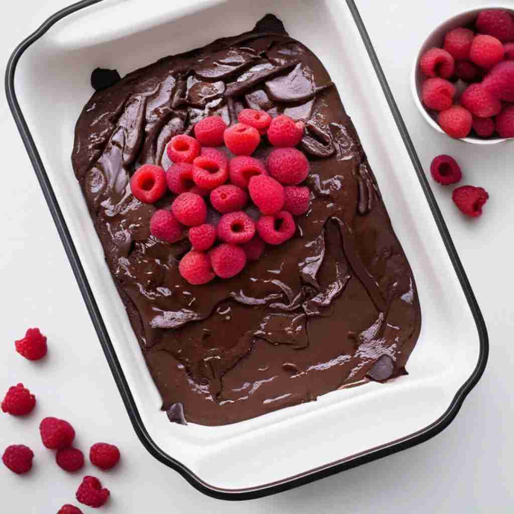 Chickpea flour brownies in a pan topped with chocolate avocado frosting and raspberries.