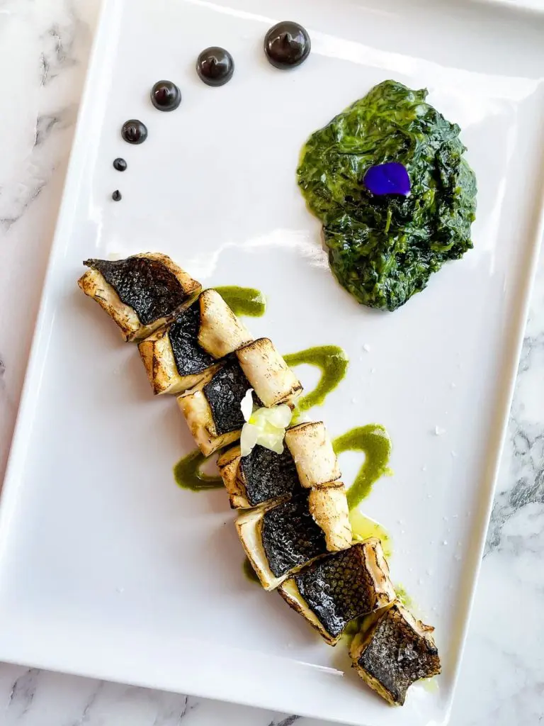 Flame-grilled wild sea bass drizzled with basil-spirulina oil, and spinach with parmesan, and dollops of sepia mayonnaise.