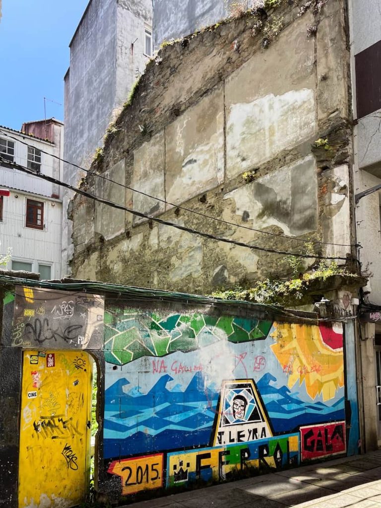 Graffiti and murals cover a wall with a yellow door that opens onto the overgrown plot of land where a house once stood.