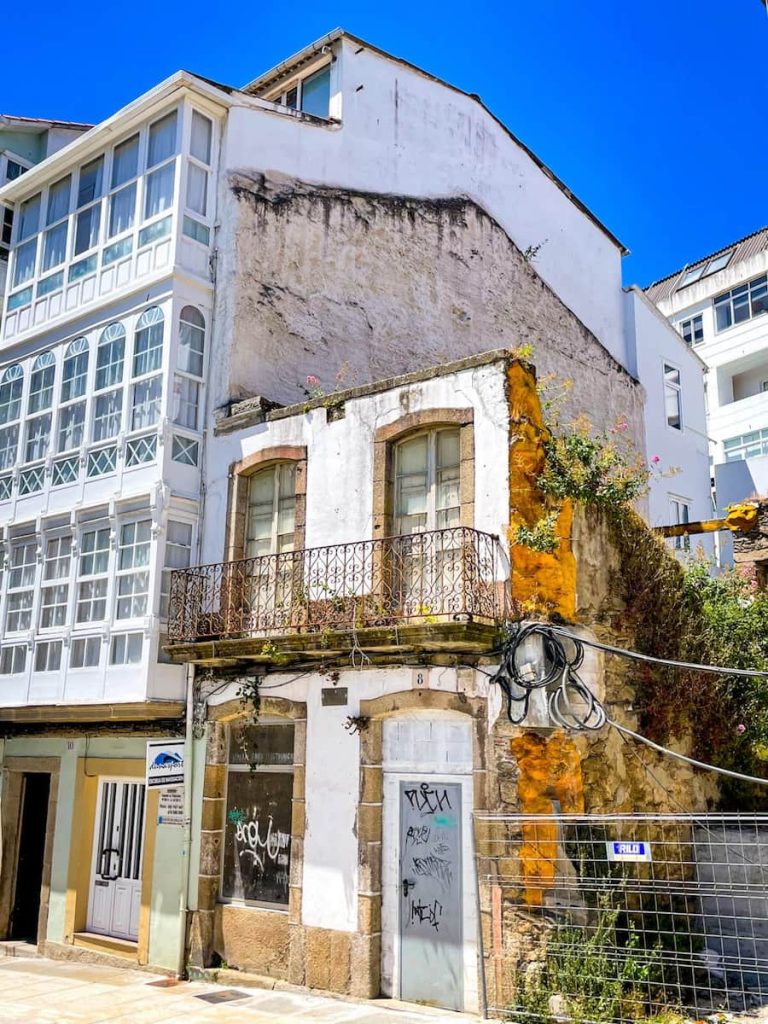 The facade of a building whose exterior has crumbled. Next to it is a traditional white house with a glassed-in balconies in Ferrol, Galicia.