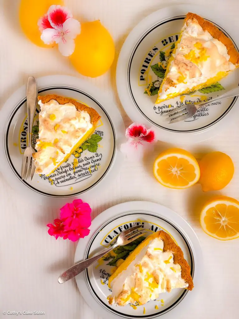 3 slices of gluten free lemon meringue pie on plates surrounded by lemons and pink flowers.