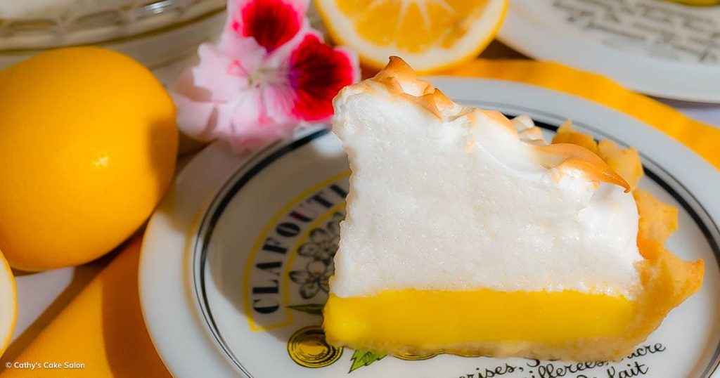 A slice of sugar free, diabetic, gluten free lemon meringue pie on a plate with lemons and a pink flower in the background.