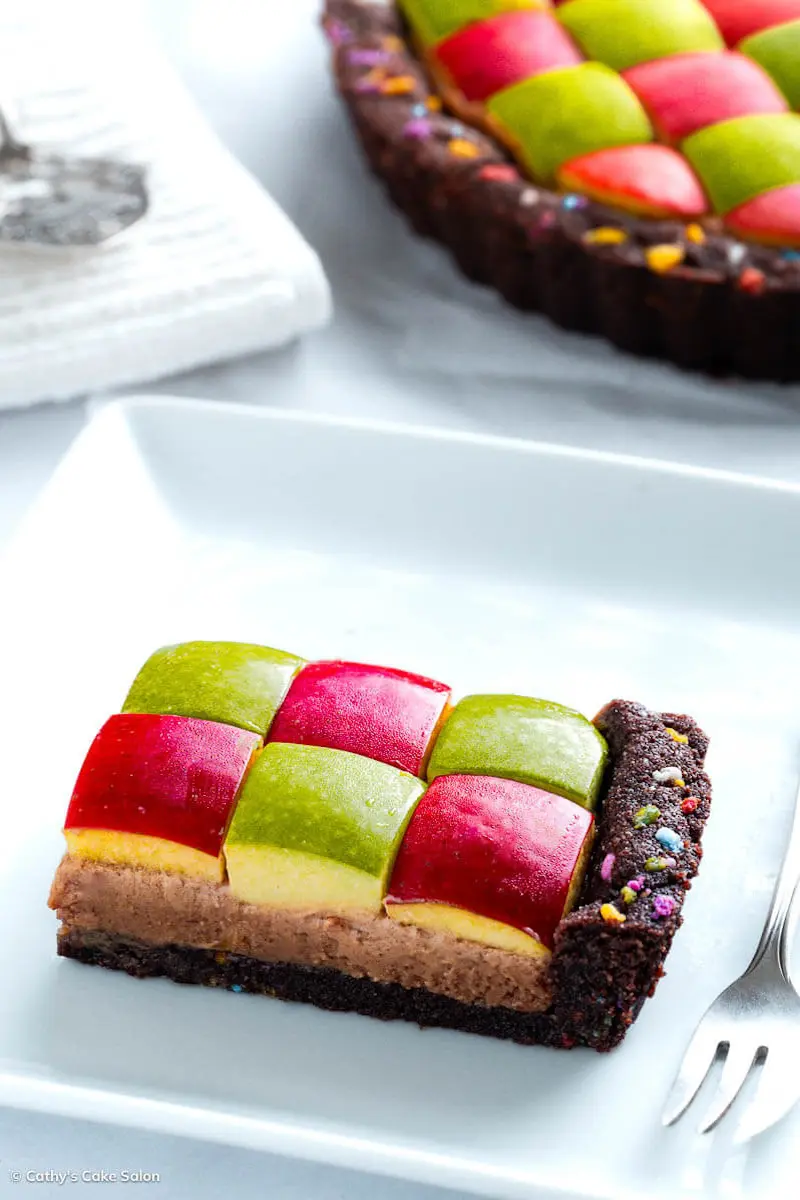 A slice of vegan peanut butter chocolate pie with apples next to the entire geometric gluten free peanut butter pie topped with red and green apple squares.
